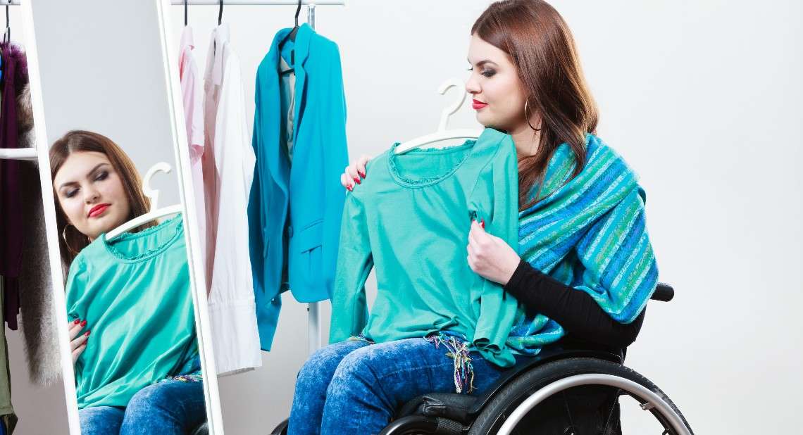 Real people, disability and handicap concept. Teen girl handicapped woman sitting on wheelchair choosing clothes in wardrobe or  looking for some clothes in shop