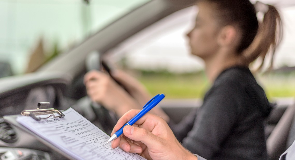 Teenage Girl Taking A Driving Lesson during bright sunny day. Closeup of a instructor man writing on a clipboard and taking notes while sitting next to teen driver.