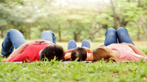 A family of three—two adults and a child—lie on their backs on a blanket in the grass, relaxing in a peaceful park. They are viewed from behind, facing the trees, enjoying a calm moment in nature together.