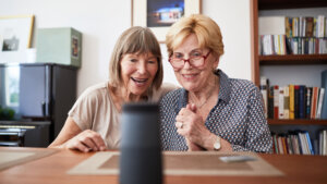 Two older women sit indoors at a table, smiling and looking at a smart speaker or voice assistant device placed in front of them. One of them wears red glasses and a patterned blouse, while the other leans in with joy. A bookshelf and framed photos are visible in the background.