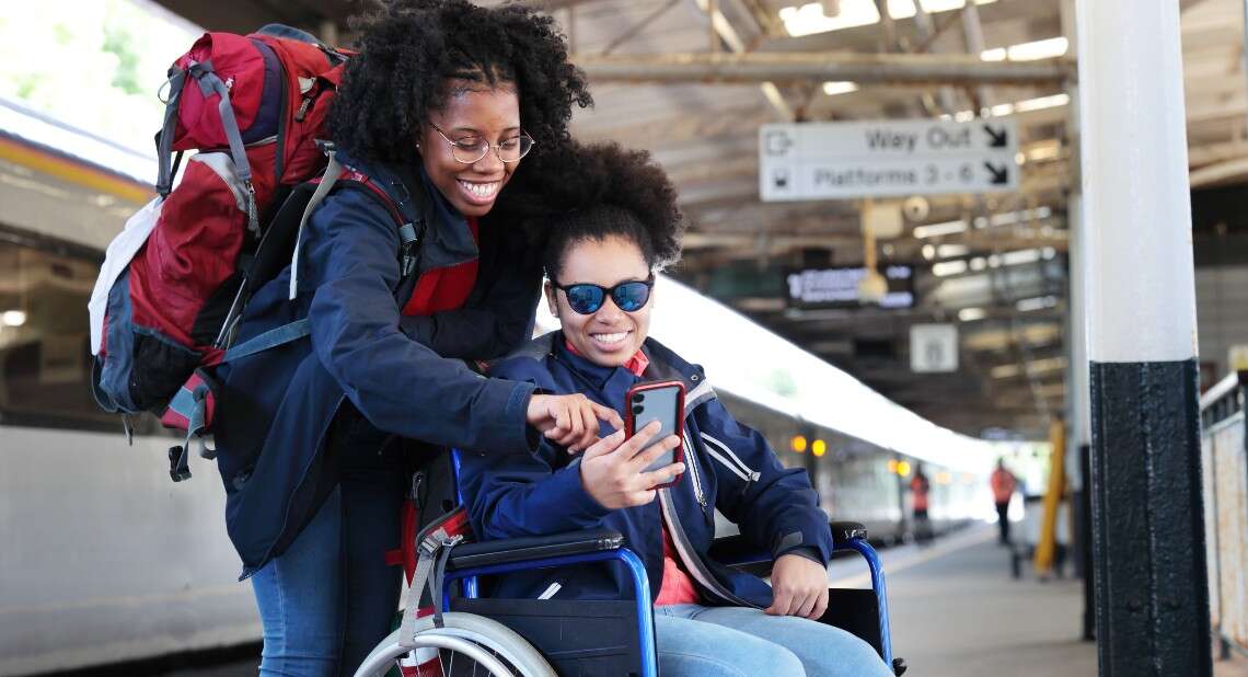 Disabled young woman in wheelchair with friend at train station looking at phone
