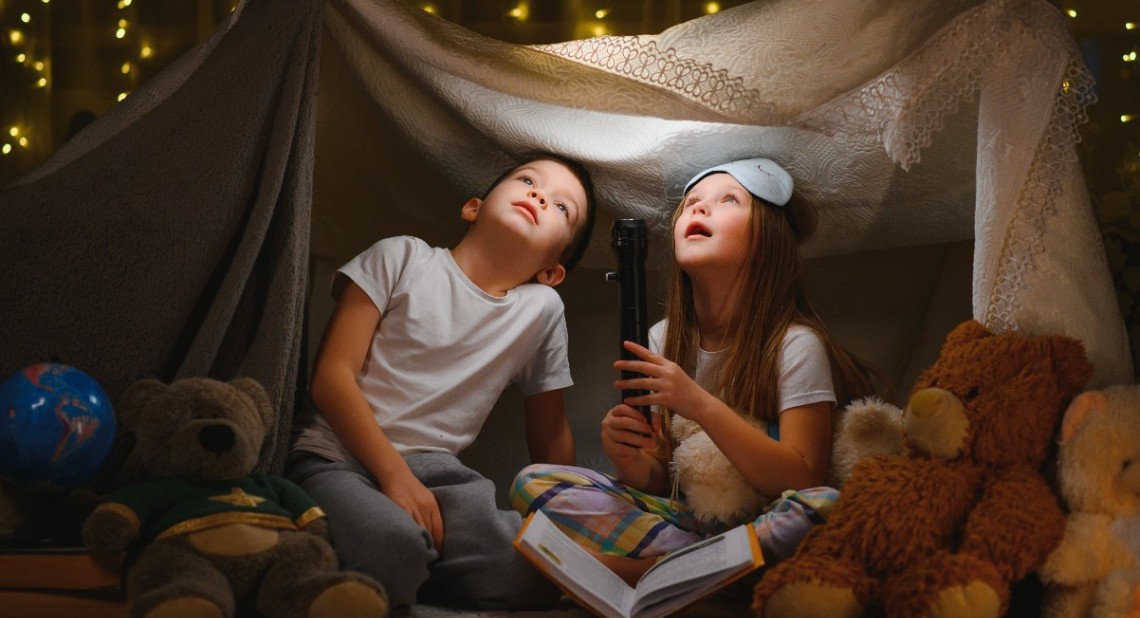 Two little child play at home in the evening to build a camping tent to read books with a flashlight and sleep inside. Concept of: game, magic, creativity, alarm systems