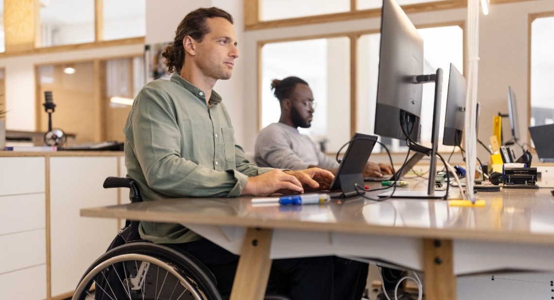 Male executive in wheelchair working on computer in the office with a colleague in background.