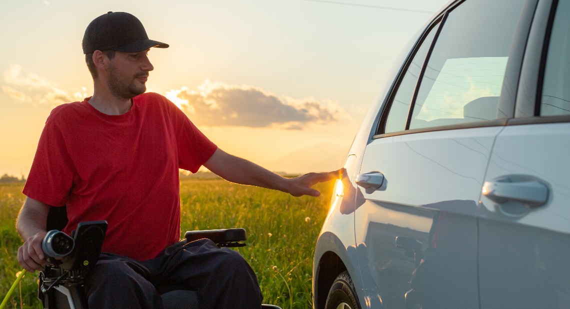 Man in wheelchair plugging in a charger in an electric car, in the background visible a nature field at sunset