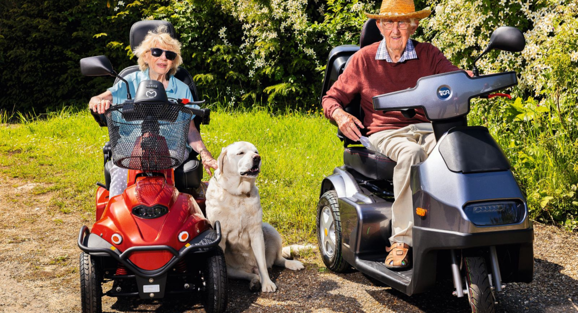 An elderly couple outdoors in their mobility scooters with their dog