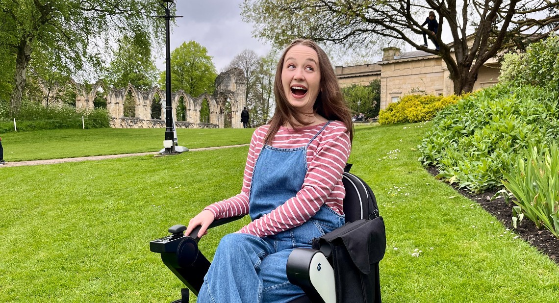 Pippa sitting in her powered wheelchair smiling, in a park