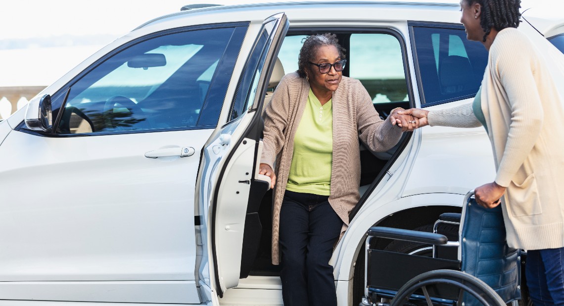 A senior woman getting into or out of a car with help from her another lady, holding her hand while she gets into her wheelchair.