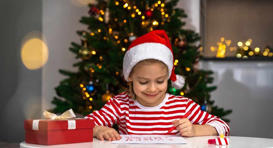 Happy little girl writes letter to Santa Claus sitting at table with gift box on background of christmas tree. New Year