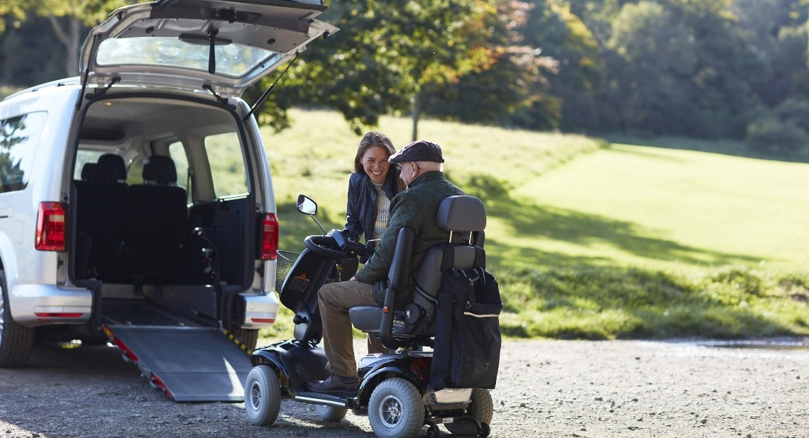 Man in a wheelchair going into his Wheelchair Accessible Vehicle with a woman by helping him