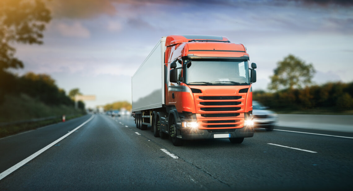 A red HGV (Heavy Goods Vehicle) truck with headlights on, driving down a highway. The truck has a large white trailer attached. The road is surrounded by greenery, with trees and a cloudy sky in the background. Other vehicles, including a car, are visible on the road, slightly blurred to indicate motion