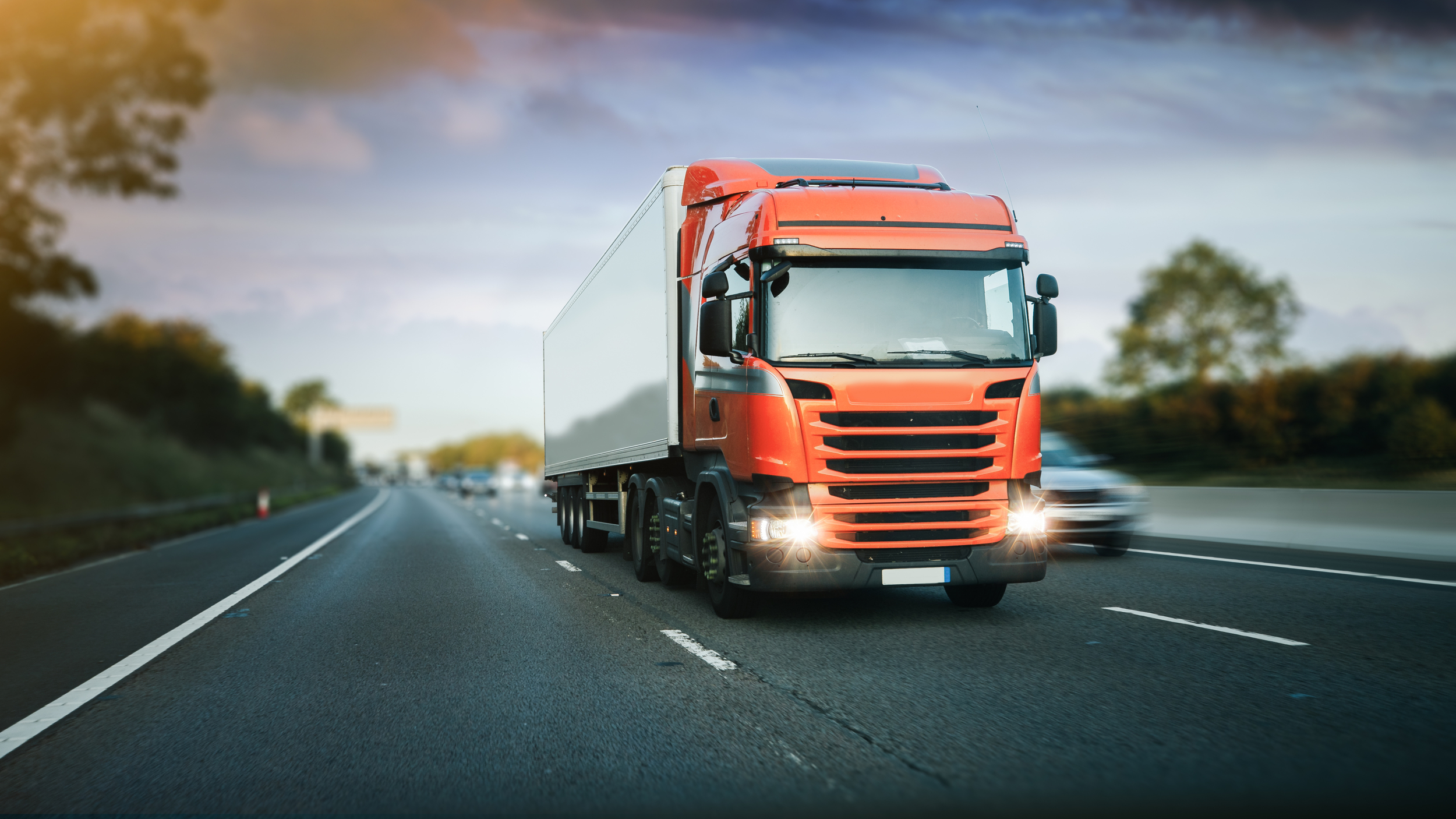 A red HGV (Heavy Goods Vehicle) truck with headlights on, driving down a highway. The truck has a large white trailer attached. The road is surrounded by greenery, with trees and a cloudy sky in the background. Other vehicles, including a car, are visible on the road, slightly blurred to indicate motion