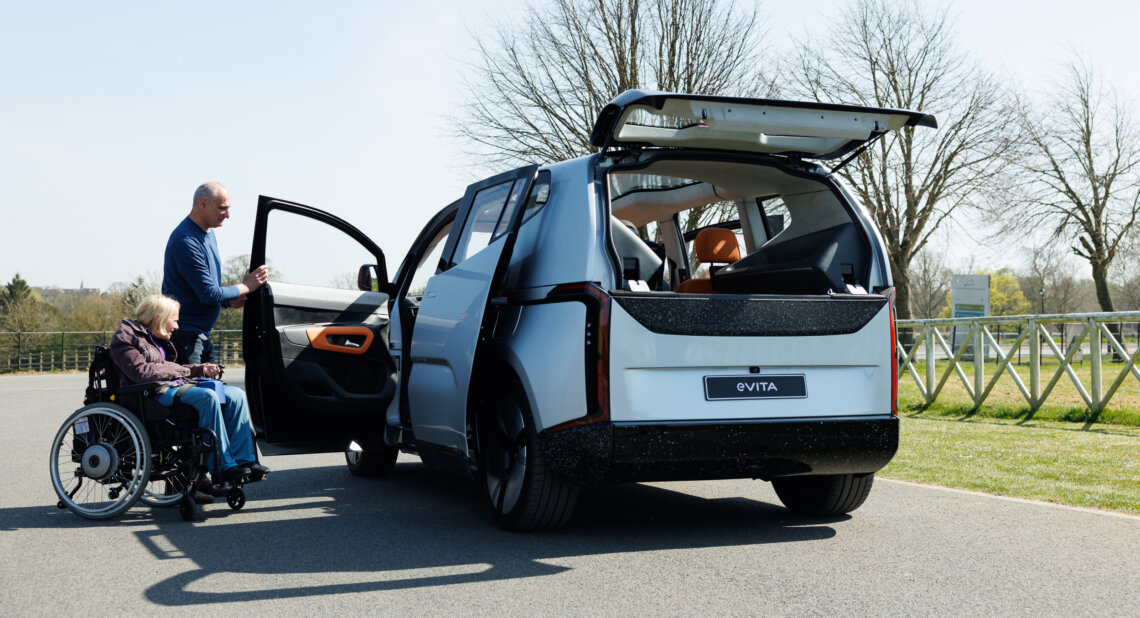 A man assists a woman in a wheelchair beside an eVITA electric vehicle parked outdoors. The car’s rear and side doors are open, showcasing its accessible design with a spacious interior and vibrant orange accents. The setting is a bright day in a park-like environment.