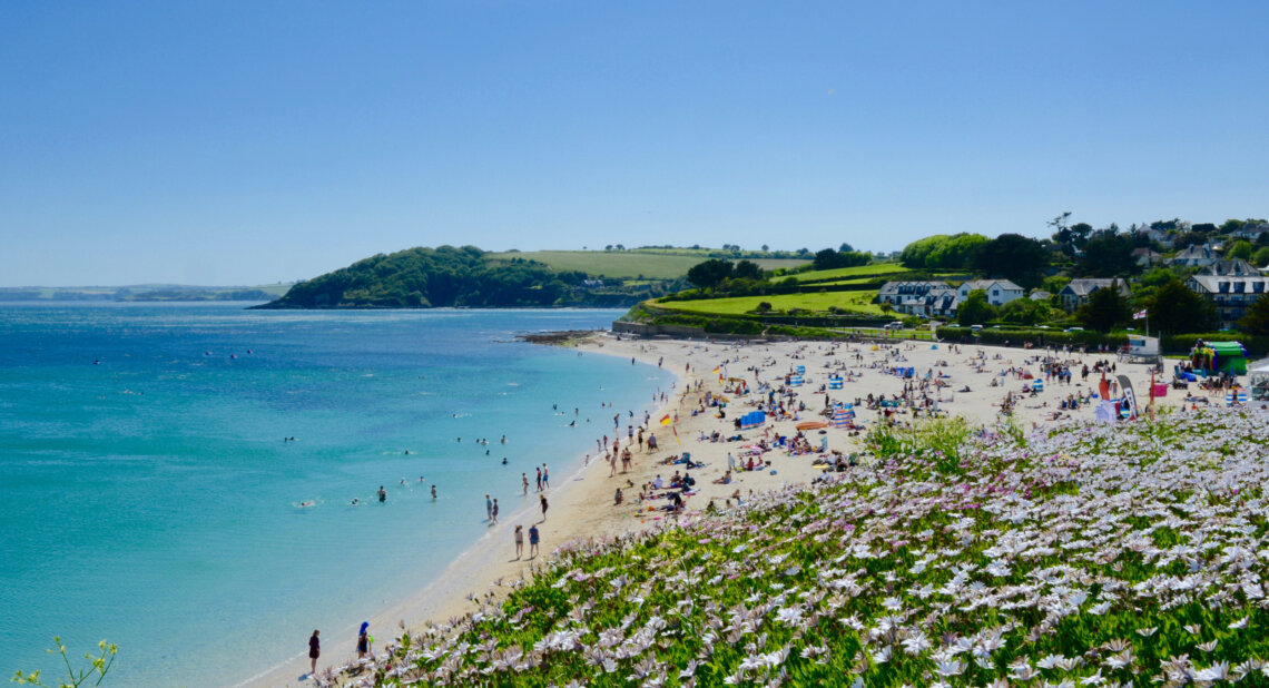 A wide sandy beach on a sunny day, with people swimming, sunbathing, and enjoying the sea. The turquoise water stretches into the distance, and a hillside covered in green fields and houses overlooks the bay. In the foreground, purple and white flowers bloom along the edge of the coast.