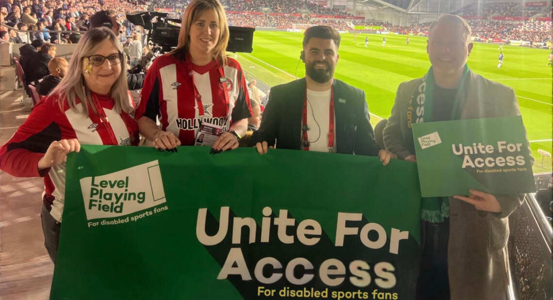 Four people, including two wearing Brentford FC shirts, stand smiling at a football stadium holding a green banner that reads “Unite For Access – For disabled sports fans” and “Level Playing Field.” The crowd and match can be seen in the background under stadium lights.