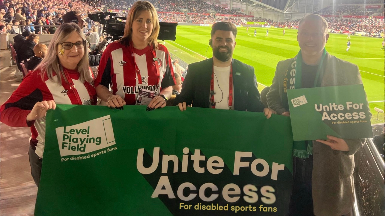 Four people, including two wearing Brentford FC shirts, stand smiling at a football stadium holding a green banner that reads “Unite For Access – For disabled sports fans” and “Level Playing Field.” The crowd and match can be seen in the background under stadium lights.