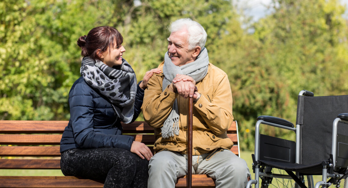 An elderly man and a younger woman sit together on a wooden park bench, smiling warmly at each other. The man leans on a walking cane and wears a tan jacket with a grey scarf, while the woman wears a dark coat and a patterned scarf. A wheelchair is positioned next to the bench, and the background features lush green trees, suggesting a peaceful outdoor setting.