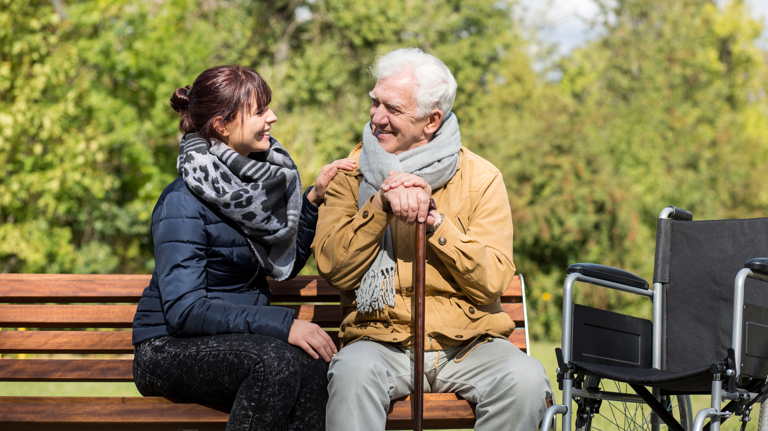 An elderly man and a younger woman sit together on a wooden park bench, smiling warmly at each other. The man leans on a walking cane and wears a tan jacket with a grey scarf, while the woman wears a dark coat and a patterned scarf. A wheelchair is positioned next to the bench, and the background features lush green trees, suggesting a peaceful outdoor setting.