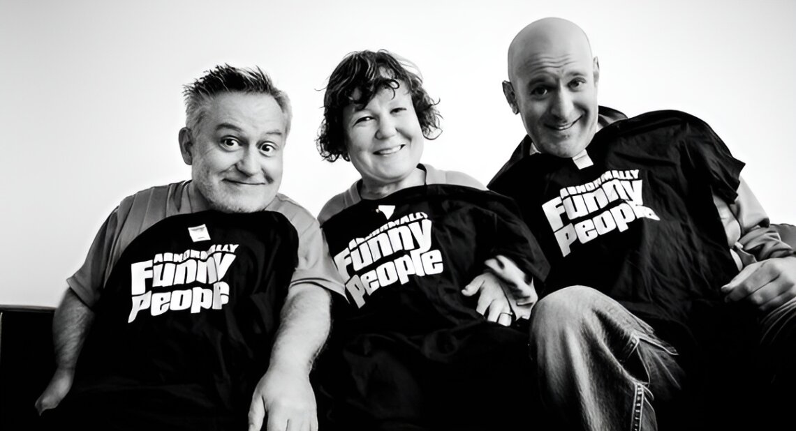 Three smiling people sit closely together, each holding or wearing a black T-shirt with the words "Abnormally Funny People" printed in bold white letters. The photo is in black and white, capturing a joyful and candid moment among the group.