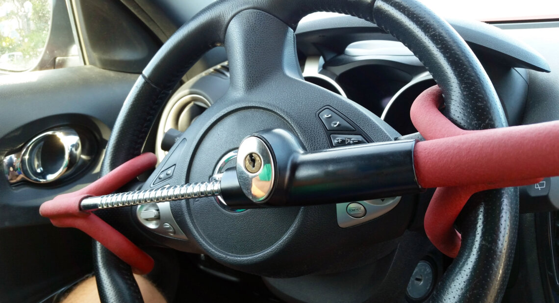 A close-up view of a car's steering wheel secured with a red and black steering wheel lock, featuring a built-in key mechanism. The anti-theft device is clamped in place to prevent the wheel from turning.