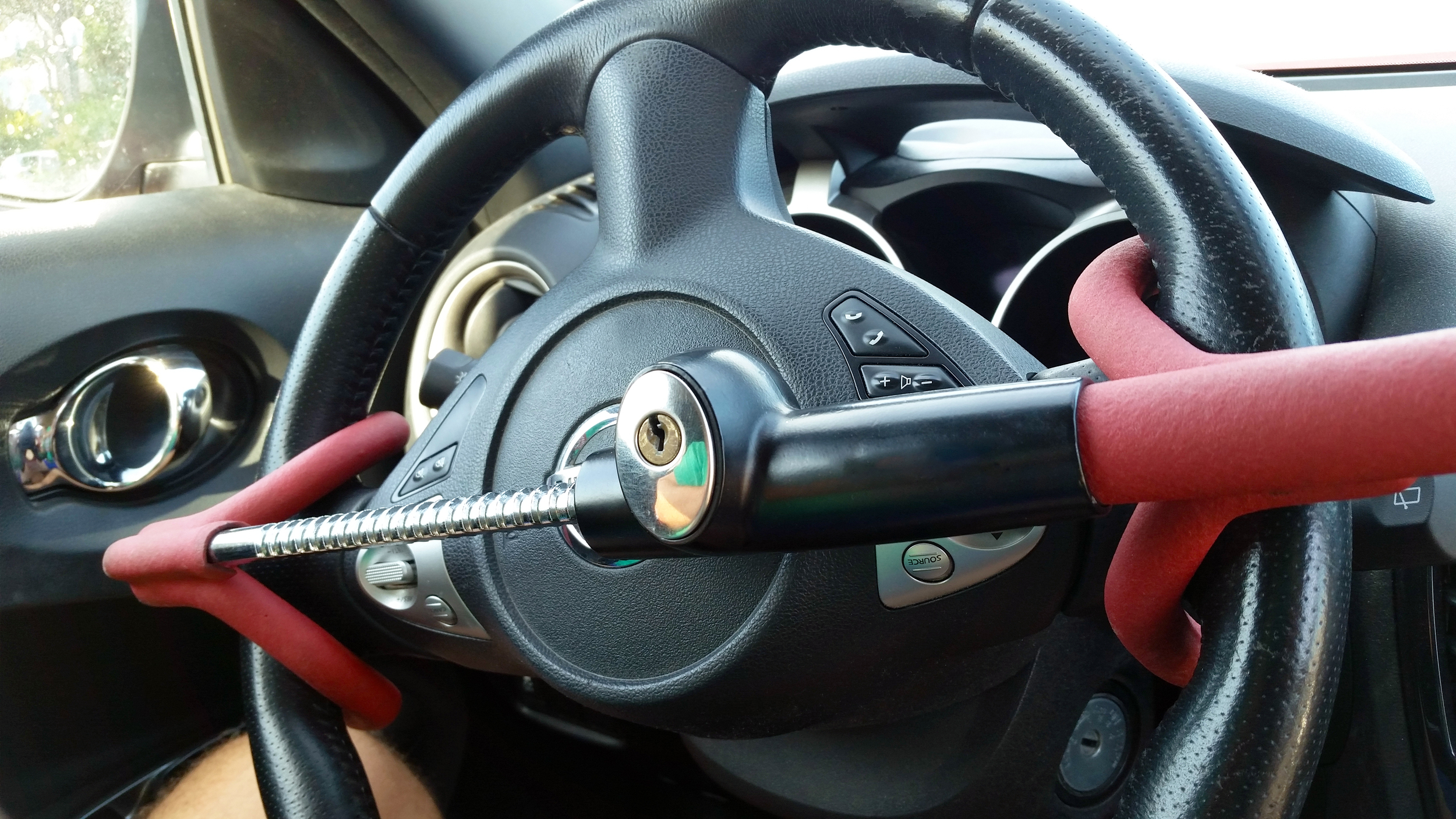 A close-up view of a car's steering wheel secured with a red and black steering wheel lock, featuring a built-in key mechanism. The anti-theft device is clamped in place to prevent the wheel from turning.