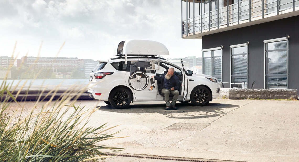 A man sits at the open side of a white SUV, preparing a mobility aid with a robotic arm that stores a wheelchair in a roof box. The vehicle is parked outside a modern building on a bright day, demonstrating advanced mobility equipment in use.