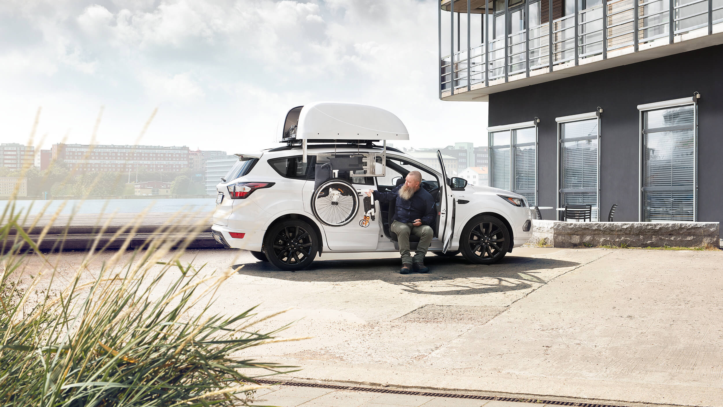 A man sits at the open side of a white SUV, preparing a mobility aid with a robotic arm that stores a wheelchair in a roof box. The vehicle is parked outside a modern building on a bright day, demonstrating advanced mobility equipment in use.