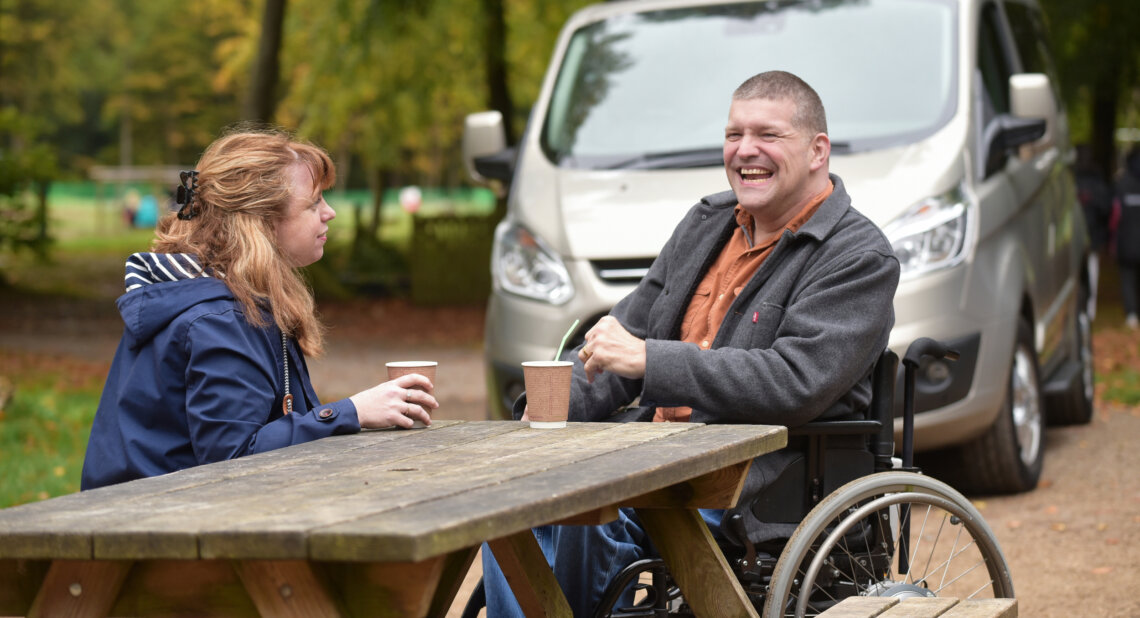 A man in a wheelchair and a woman sit across from each other at a wooden picnic table in a park, enjoying hot drinks and smiling. A wheelchair-accessible van is parked behind them, and the scene is set in a leafy outdoor area during autumn.