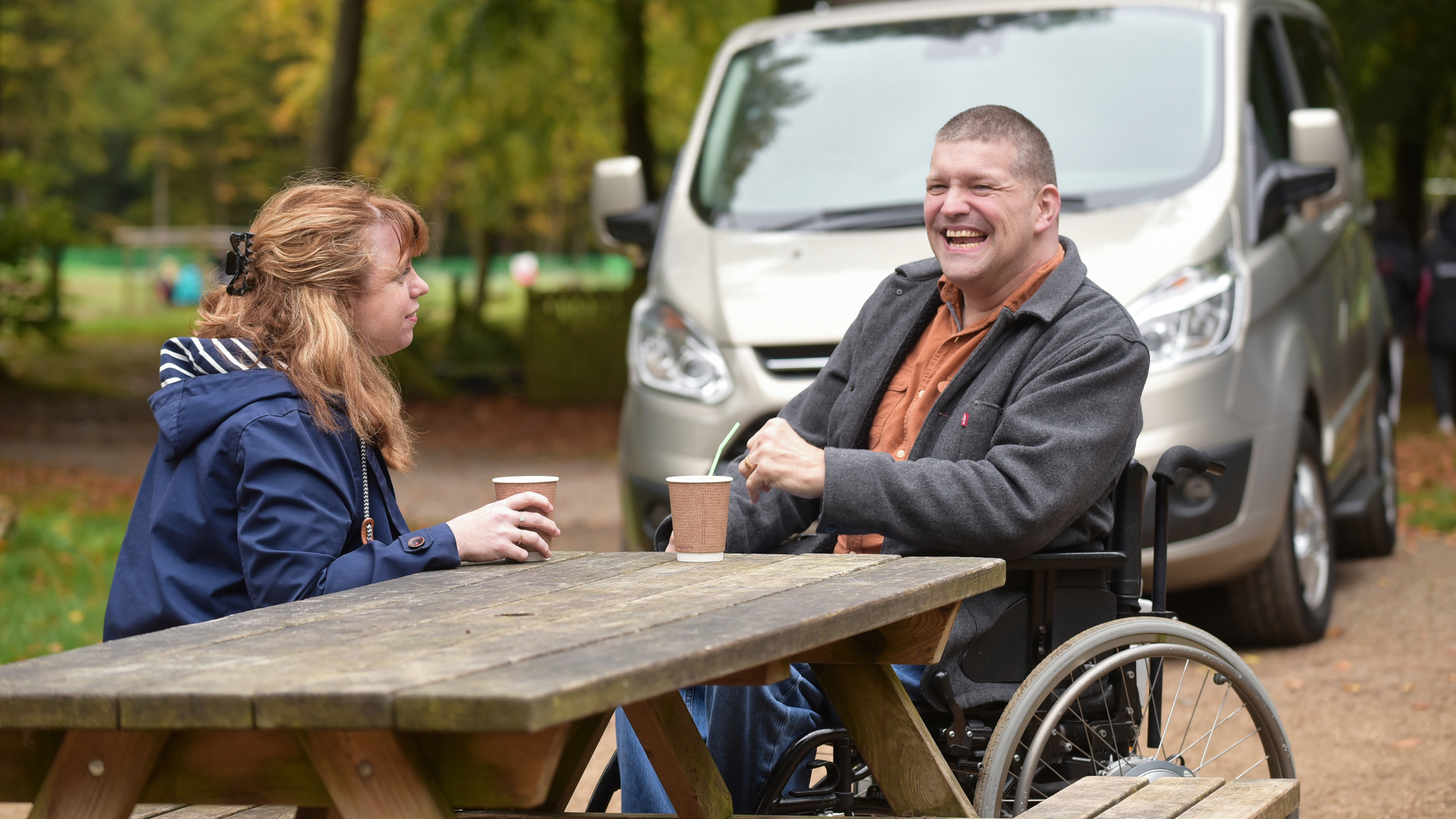 A man in a wheelchair and a woman sit across from each other at a wooden picnic table in a park, enjoying hot drinks and smiling. A wheelchair-accessible van is parked behind them, and the scene is set in a leafy outdoor area during autumn.