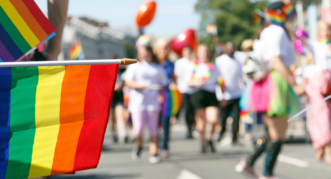 Close-up of a rainbow Pride flag being waved at a lively outdoor Pride parade. In the blurred background, people dressed in colourful clothing and accessories walk together, some holding flags and balloons, celebrating LGBTQ+ pride and inclusivity.