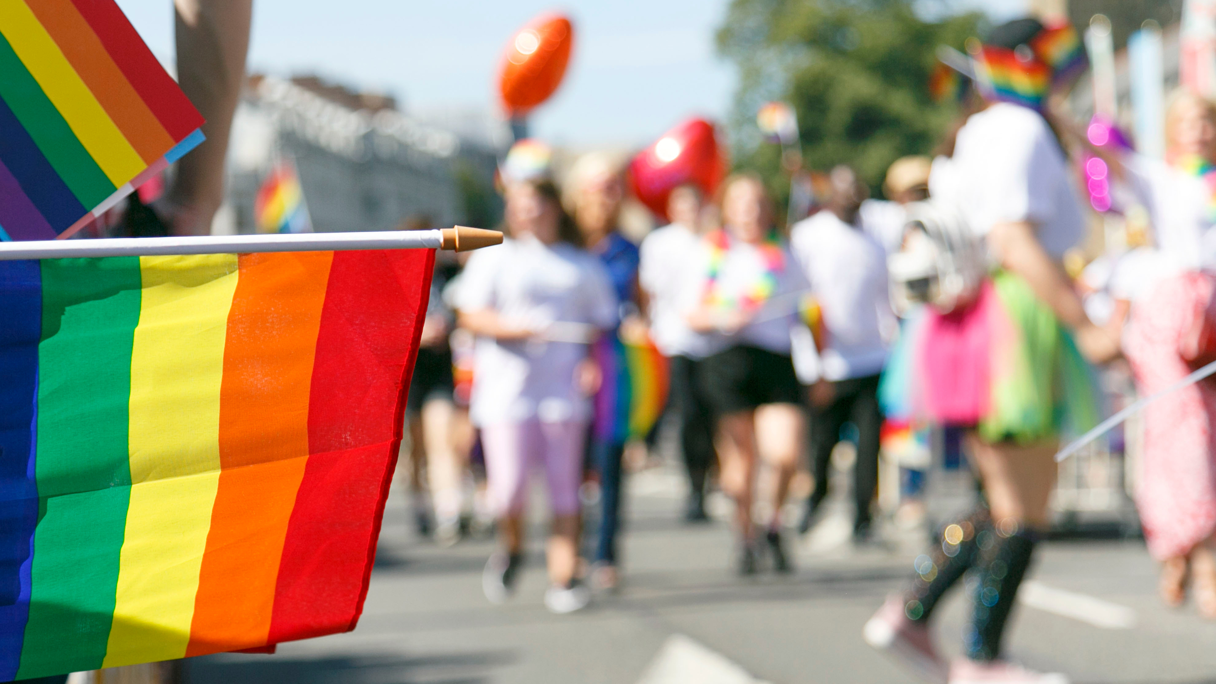 Close-up of a rainbow Pride flag being waved at a lively outdoor Pride parade. In the blurred background, people dressed in colourful clothing and accessories walk together, some holding flags and balloons, celebrating LGBTQ+ pride and inclusivity.
