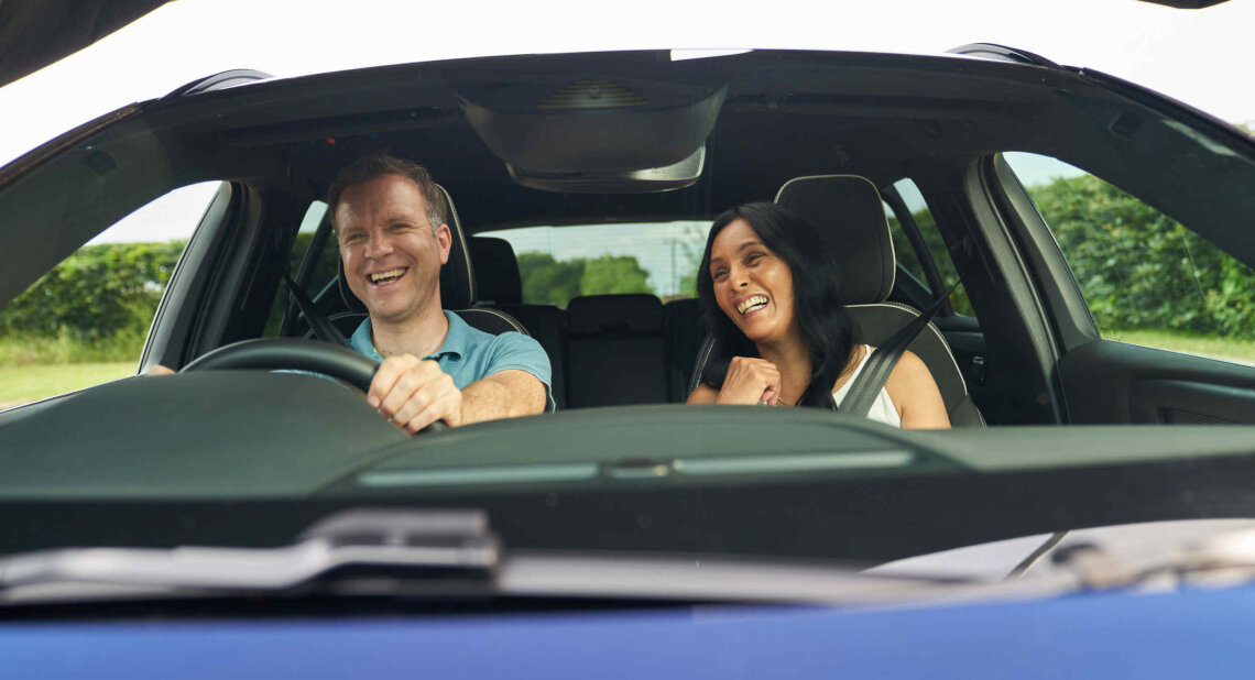 A man and woman sit in the front seats of a car, both smiling and laughing as they enjoy a drive together. The photo is taken from outside through the windshield, capturing their cheerful expressions and the green landscape visible through the windows.
