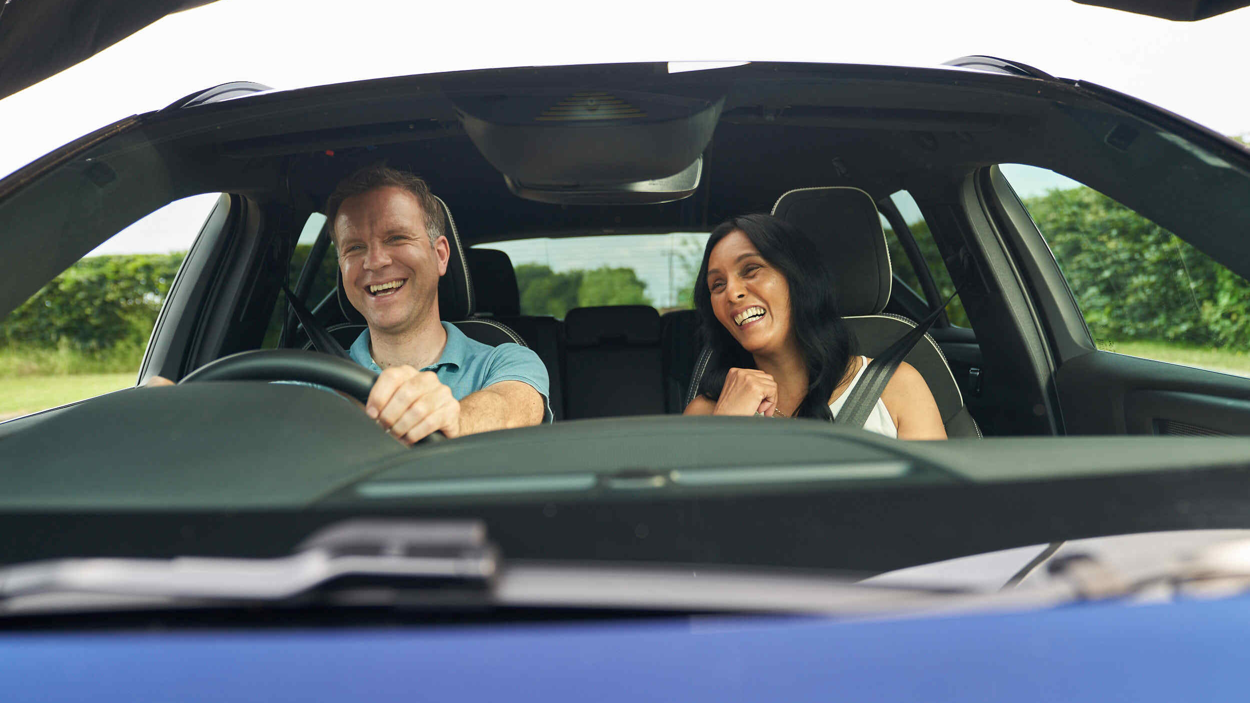 A man and woman sit in the front seats of a car, both smiling and laughing as they enjoy a drive together. The photo is taken from outside through the windshield, capturing their cheerful expressions and the green landscape visible through the windows.