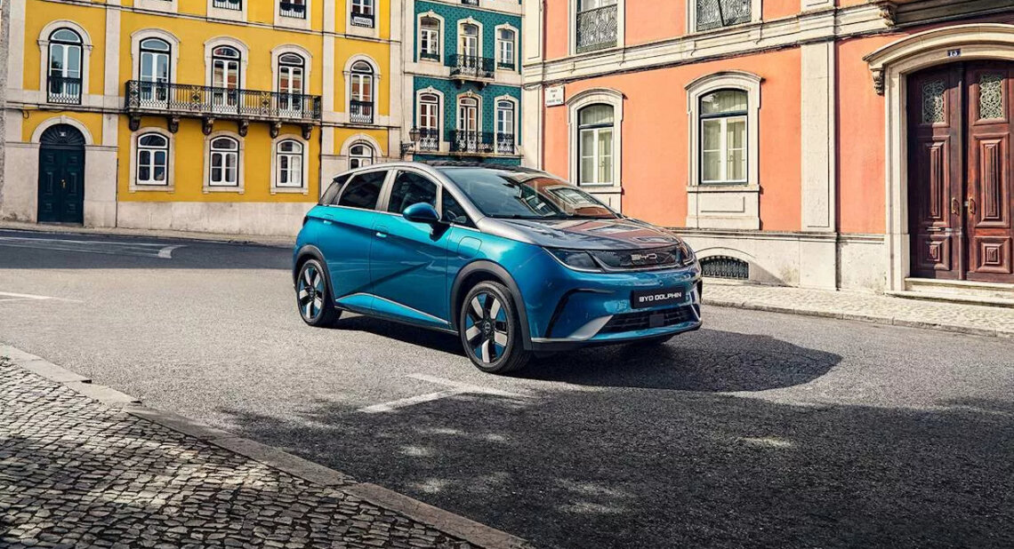A blue BYD Dolphin electric car is parked on a quiet, sunny street lined with colourful European-style buildings in yellow, teal, and peach tones. The vehicle is positioned at an angle, showing its modern and compact design.