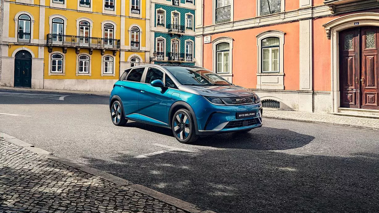 A blue BYD Dolphin electric car is parked on a quiet, sunny street lined with colourful European-style buildings in yellow, teal, and peach tones. The vehicle is positioned at an angle, showing its modern and compact design.