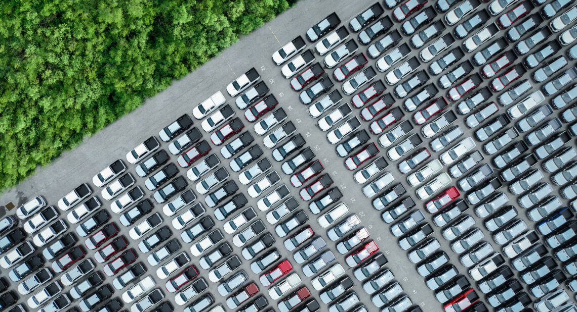 Aerial view of a large car park filled with rows of white, black, silver, and red vehicles, bordered by dense green trees on one side.