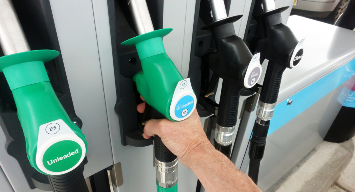 A person’s hand grips a green fuel pump nozzle labeled E5 unleaded at a petrol station, next to black diesel nozzles labeled B7.