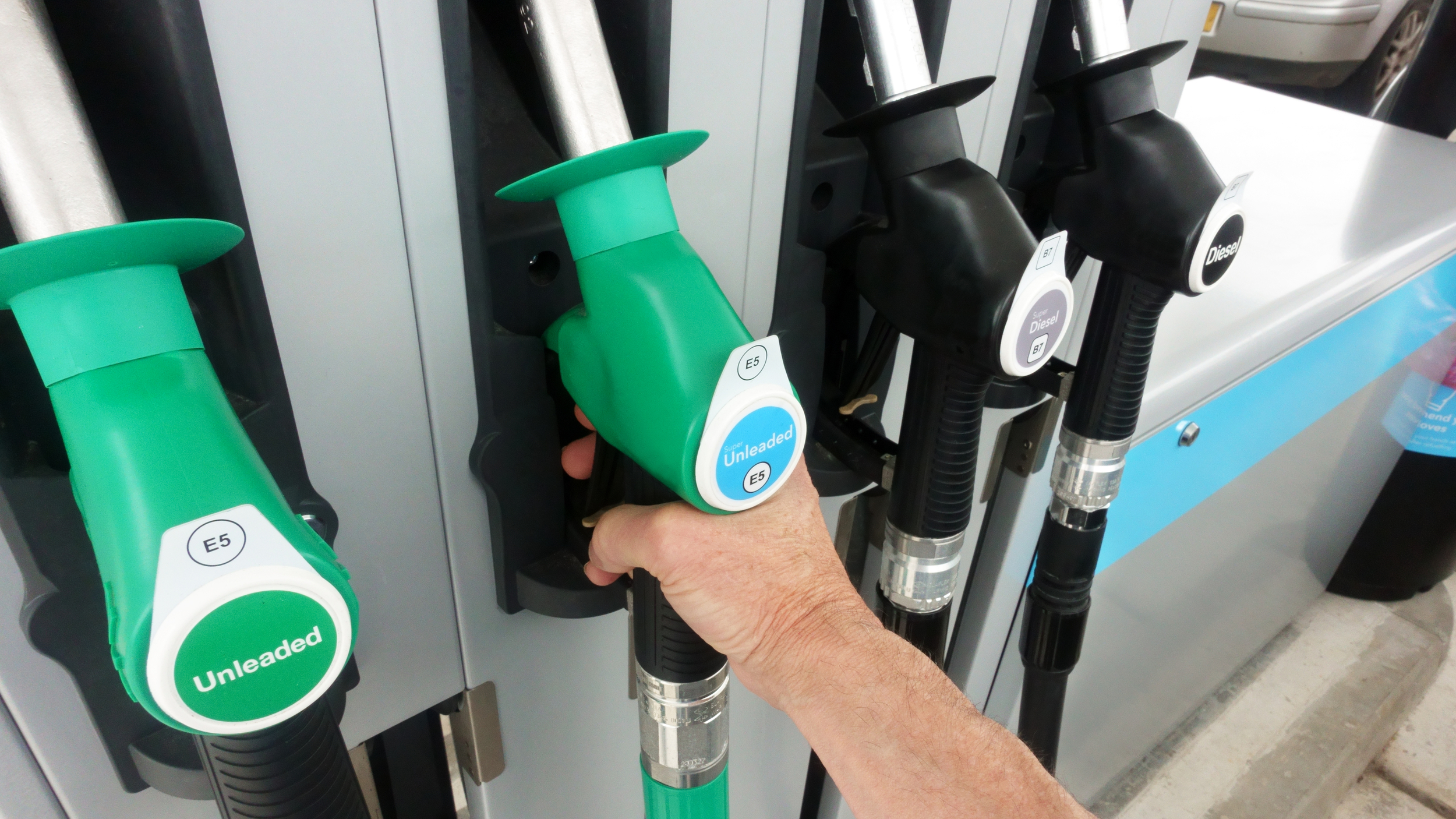 A person’s hand grips a green fuel pump nozzle labeled E5 unleaded at a petrol station, next to black diesel nozzles labeled B7.