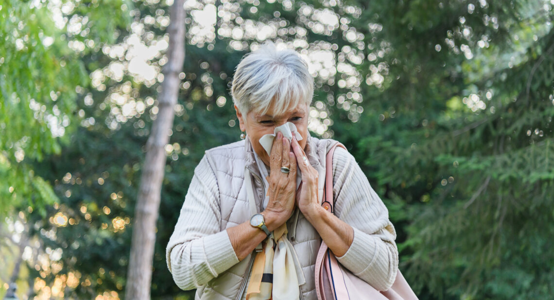 An older woman stands outdoors surrounded by trees, holding a tissue to her nose as she sneezes. She wears a light quilted vest and scarf, and carries a pink handbag over her shoulder, suggesting she is experiencing hay fever or seasonal allergies.