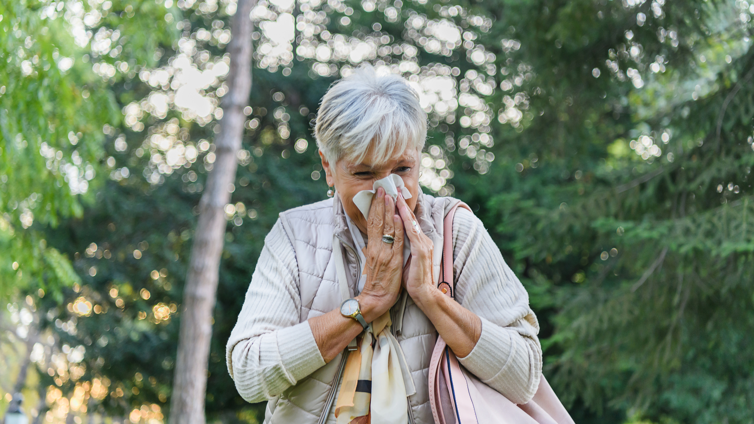 An older woman stands outdoors surrounded by trees, holding a tissue to her nose as she sneezes. She wears a light quilted vest and scarf, and carries a pink handbag over her shoulder, suggesting she is experiencing hay fever or seasonal allergies.