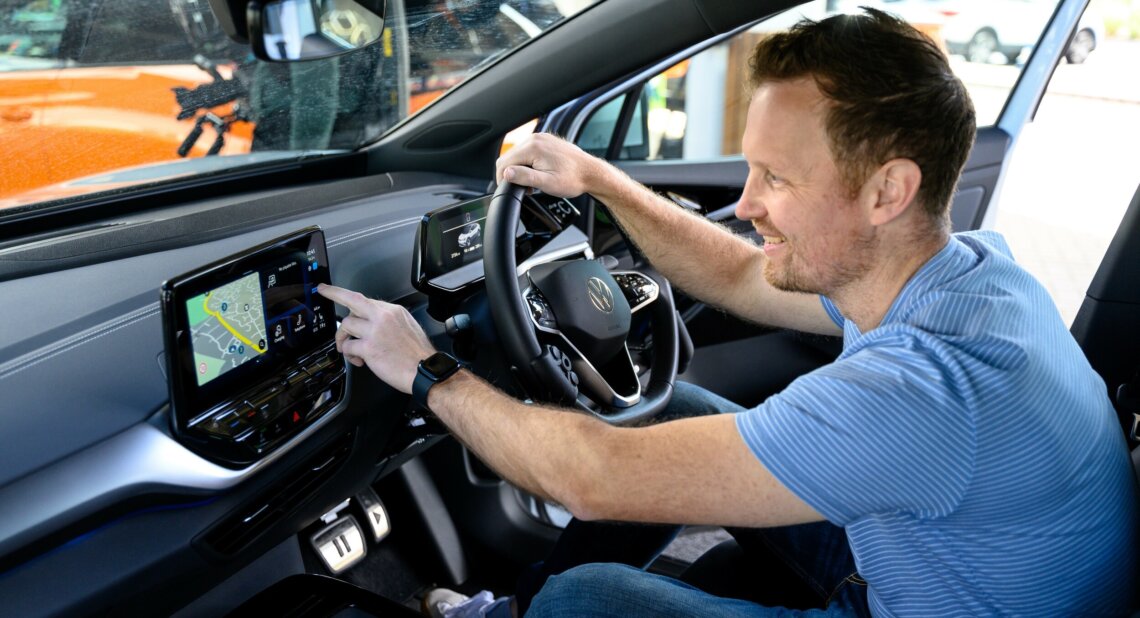 James Hunt smiles as he sits in the driver’s seat of an electric vehicle, interacting with the touchscreen navigation and infotainment system on the dashboard.