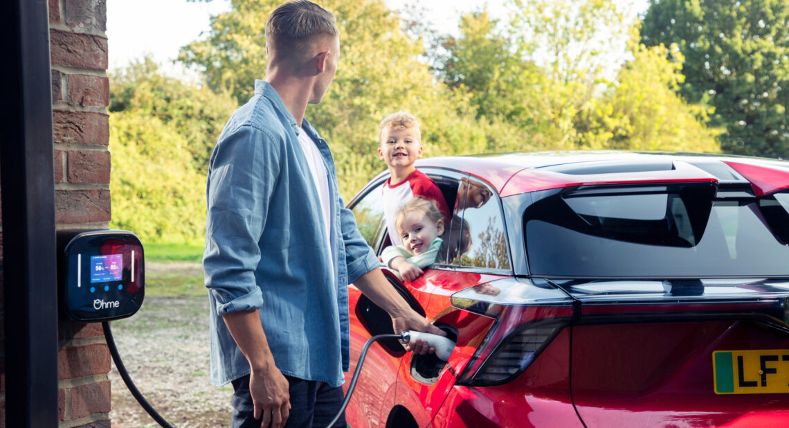 A man charges a red electric vehicle at home using an Ohme wall charger mounted on a brick wall. Two young children smile and look out from the rear windows of the car, enjoying the moment. The setting is a sunny day with greenery in the background.