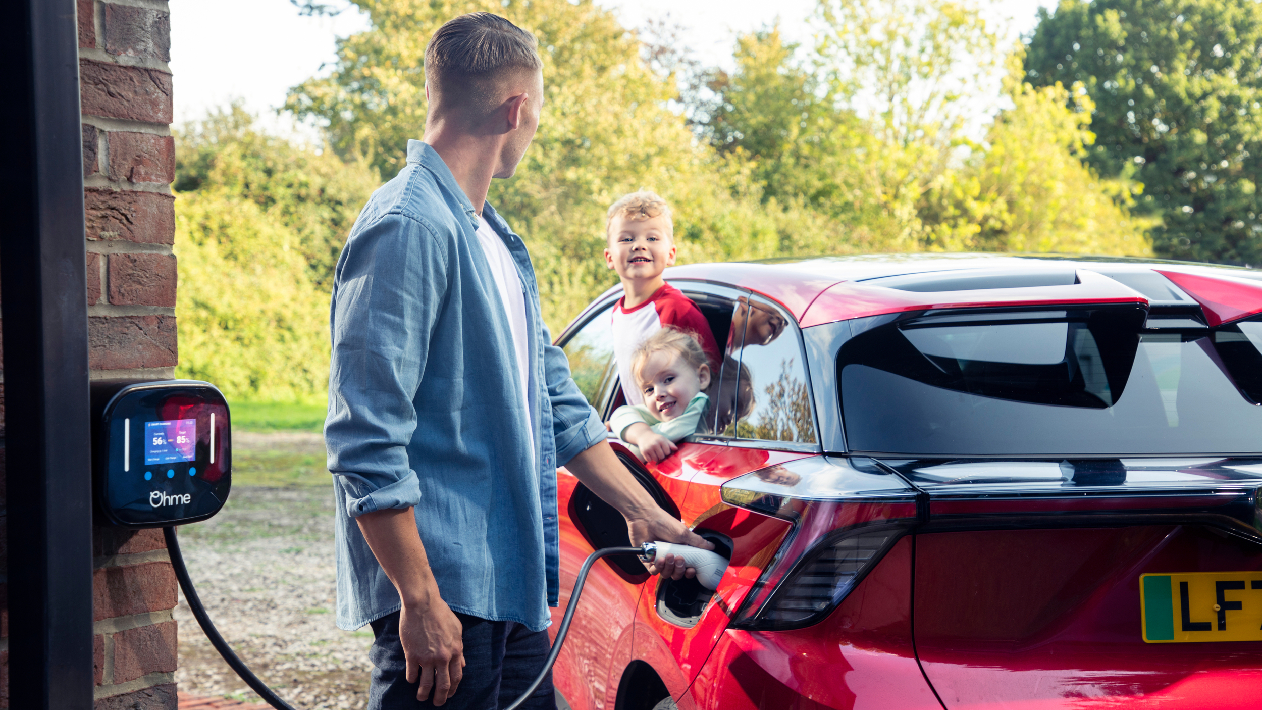 A man charges a red electric vehicle at home using an Ohme wall charger mounted on a brick wall. Two young children smile and look out from the rear windows of the car, enjoying the moment. The setting is a sunny day with greenery in the background.