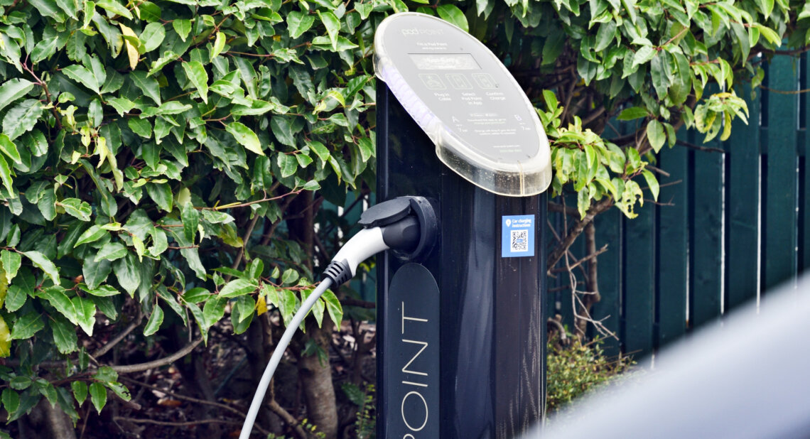 A Pod Point electric vehicle charging station is installed outdoors next to a green hedge, with a charging cable plugged in and a QR code visible on the unit.