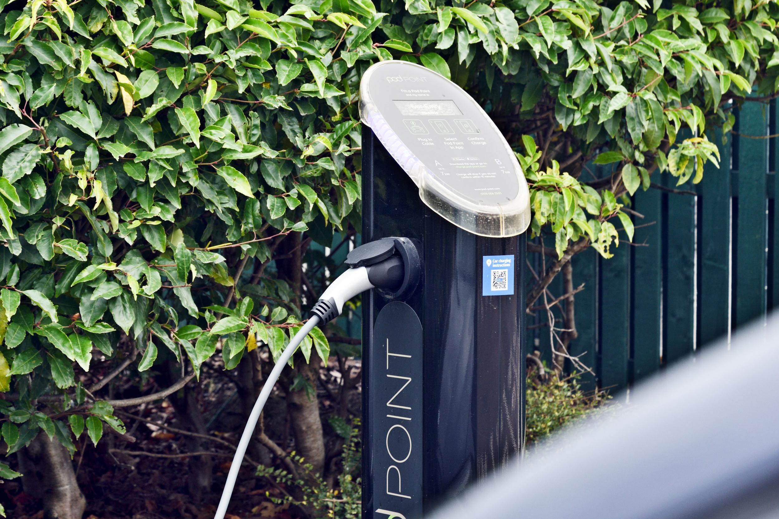 A Pod Point electric vehicle charging station is installed outdoors next to a green hedge, with a charging cable plugged in and a QR code visible on the unit.