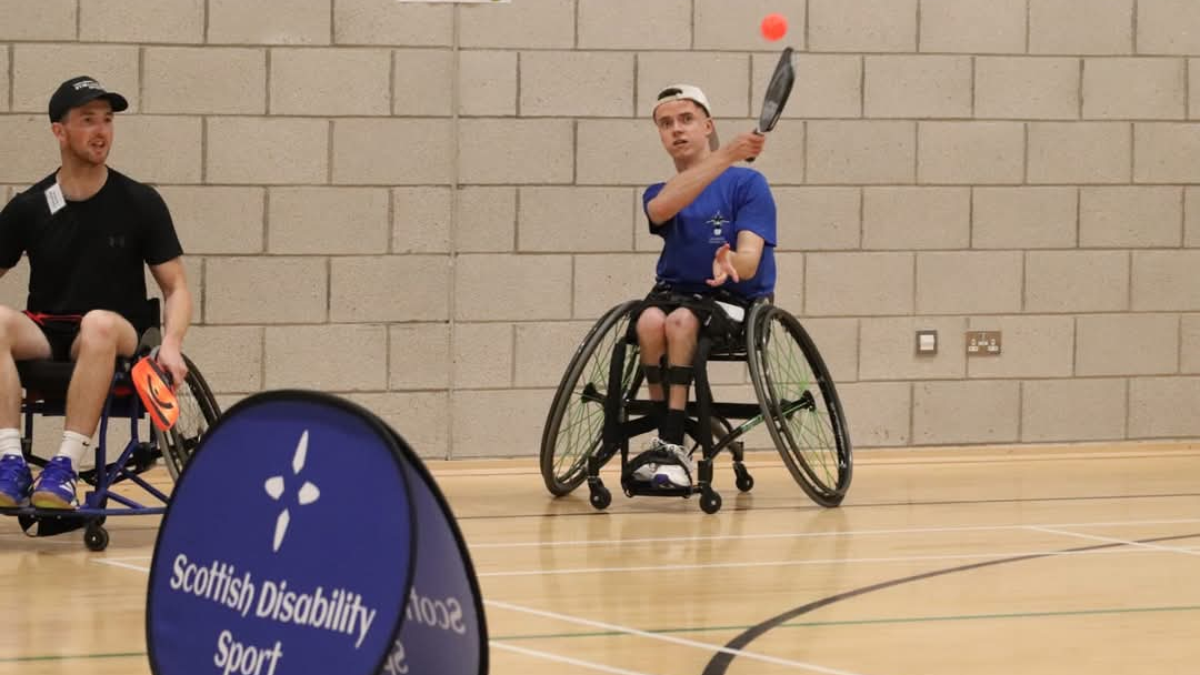 Two athletes in wheelchairs play a game of tennis indoors, with one of them mid-swing hitting an orange ball. A Scottish Disability Sport banner is visible in the foreground.