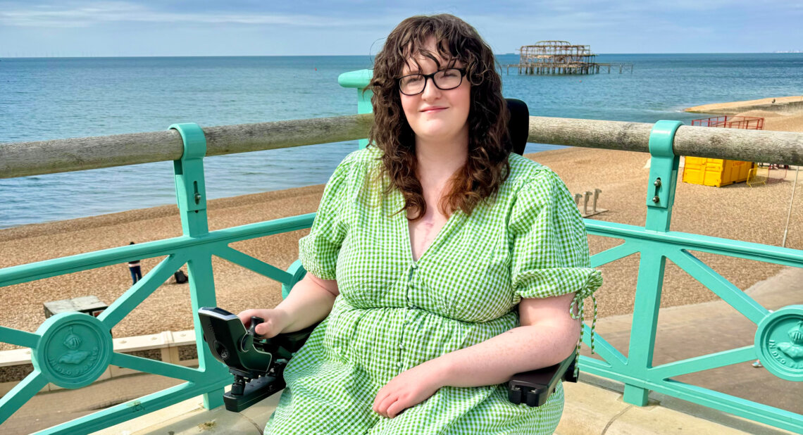 Shona Louise, a wheelchair user, smiles while seated along a seaside promenade with a turquoise railing. She is wearing a green checkered dress, with the calm ocean and Brighton's historic West Pier ruins visible in the background on a sunny day.