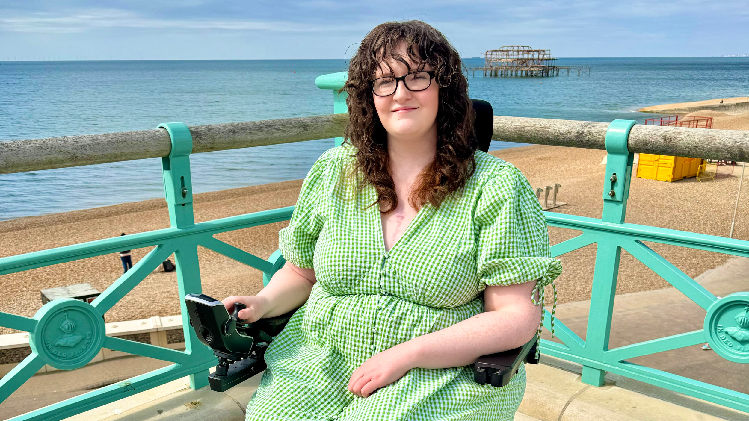 Shona Louise, a wheelchair user, smiles while seated along a seaside promenade with a turquoise railing. She is wearing a green checkered dress, with the calm ocean and Brighton's historic West Pier ruins visible in the background on a sunny day.
