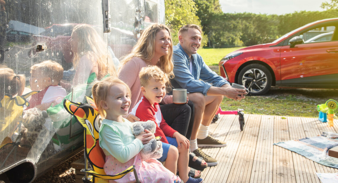A smiling family of four sits outside a shiny caravan on a sunny day. Two young children sit on colourful chairs, while the parents sit beside them holding mugs. A red car is parked in the background on a grassy campsite.