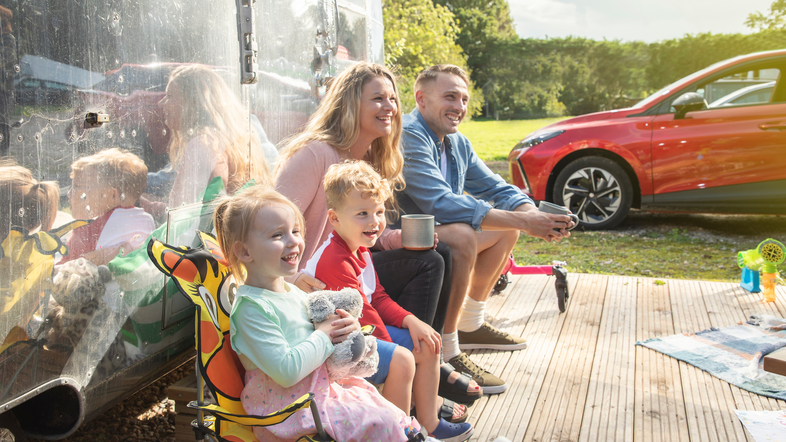 A smiling family of four sits outside a shiny caravan on a sunny day. Two young children sit on colourful chairs, while the parents sit beside them holding mugs. A red car is parked in the background on a grassy campsite.