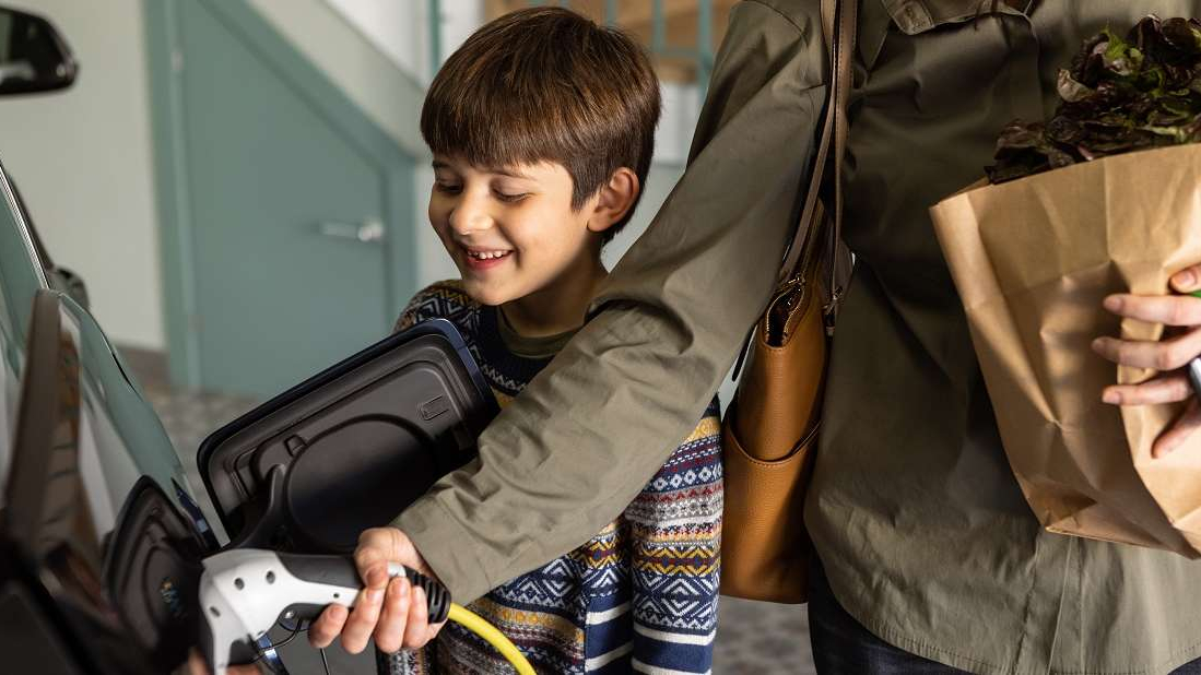 A young boy smiles as he watches an adult plug a charging cable into an electric vehicle. The adult is holding a paper grocery bag filled with produce and has a handbag over one shoulder. The scene takes place in a home garage or parking area.